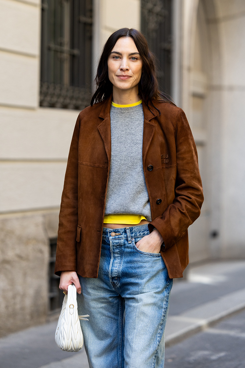 Alexa Chung in Milan wearing a Miu Miu brown suede jacket with a gray sweater over a yellow T-shirt, baggy jeans and ballet flats.