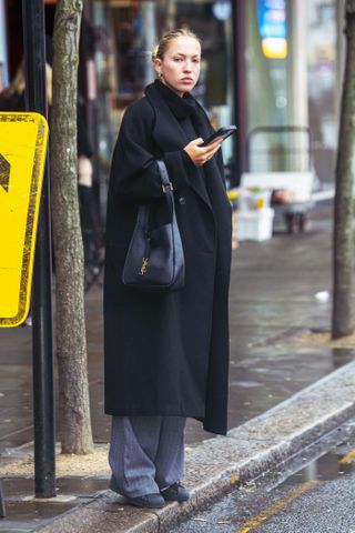 A photo of Lila Moss pairing gray pinstripe pants with black Adidas Superstar sneakers, a black coat, and a black turtleneck sweater.