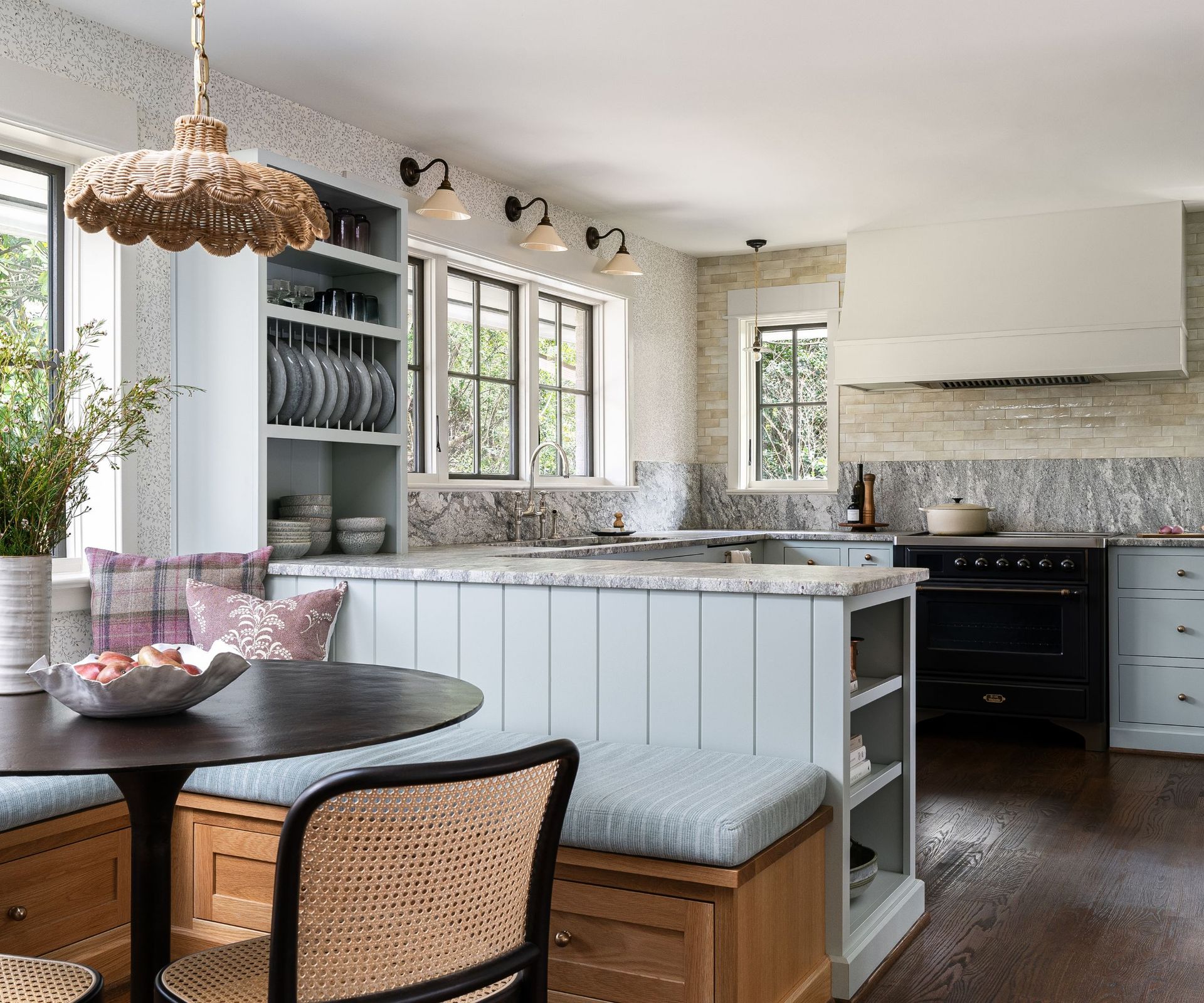 A blue peninsula kitchen with grey and white marble countertops and backsplash. Layered lighting has been introduced with pendant lights beside the range, wall lights above the window, and a rattan pendant above the table in the dining nook
