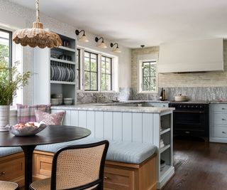 A blue peninsula kitchen with grey and white marble countertops and backsplash. Layered lighting has been introduced with pendant lights beside the range, wall lights above the window, and a rattan pendant above the table in the dining nook