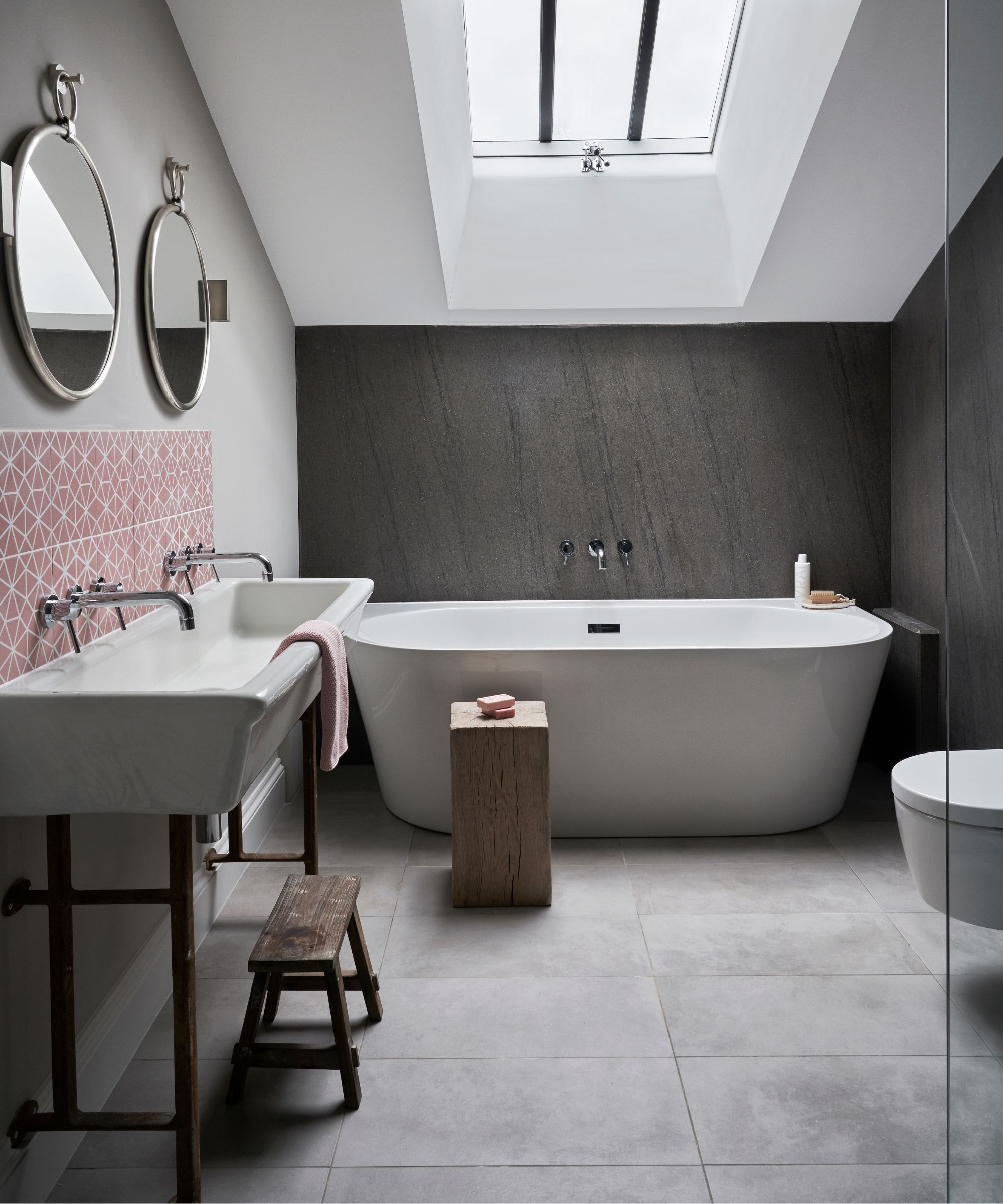 A neutral attic bathroom space with a white ceiling, wooden walls, white bathtub, wooden stools, white sink, red and white patterned backsplash and two circular mirrors above.