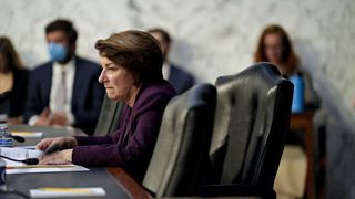 A side-on photo of Senator Amy Klobuchar sat at a desk with people behind sat by a wall
