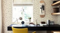 a room with black detailed wallpaper, a black desk, a yellow chair, a skateboard hanging on the wall, and various objects spread out across the desk