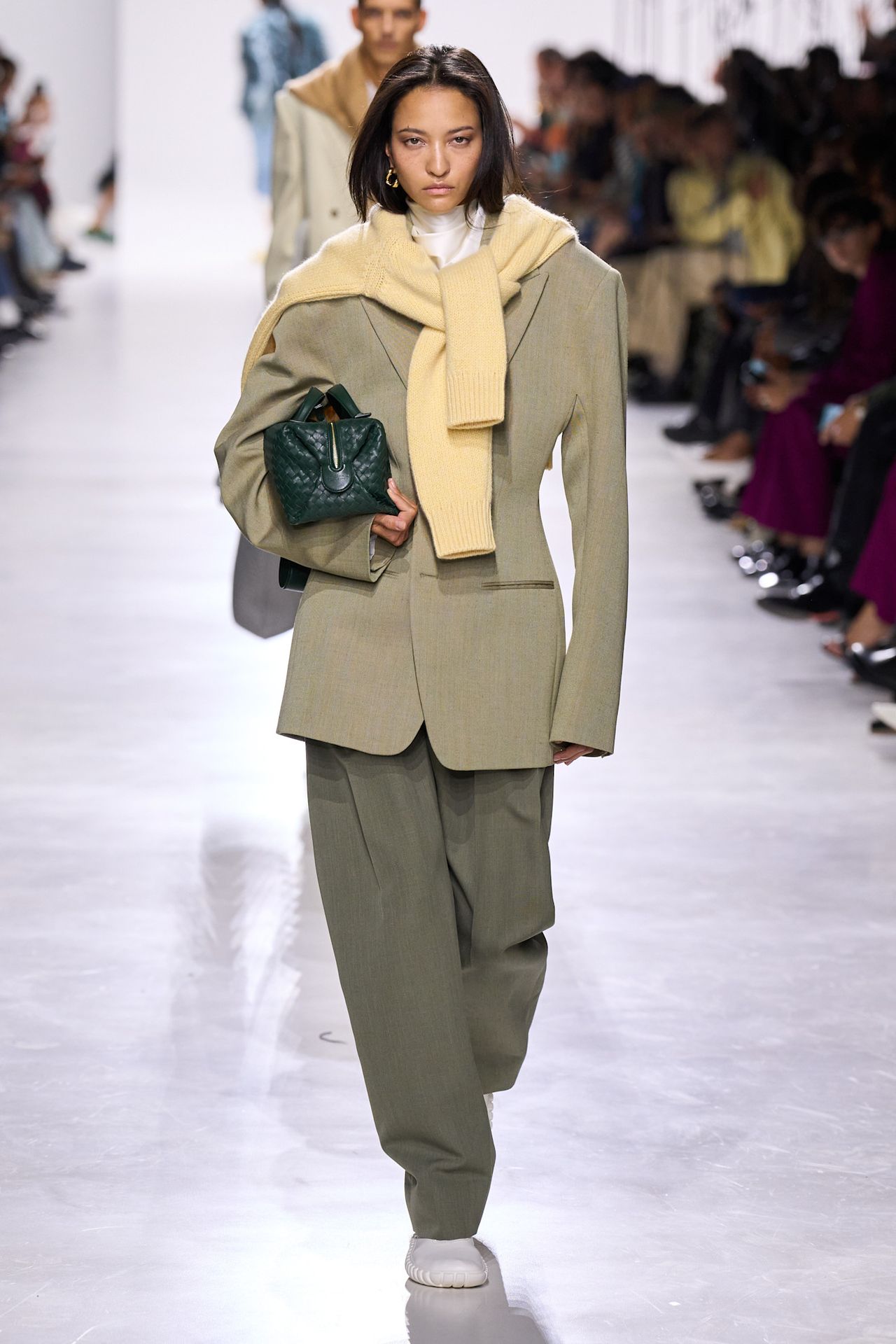 Layering staples for spring 2026 is shown in a photo from Bottega Veneta's runway show, with a model walking while wearing a yellow sweater tied over the shoulders with a white turtleneck layered underneath a sage green blazer styled with sage green trousers, white flats, and a green clutch bag