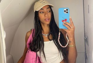 Dutch fashion influencer Amaka Hamelijnck taking a selfie on an airplane wearing a white tank, a baseball hat, denim shorts, and a pink tote bag.