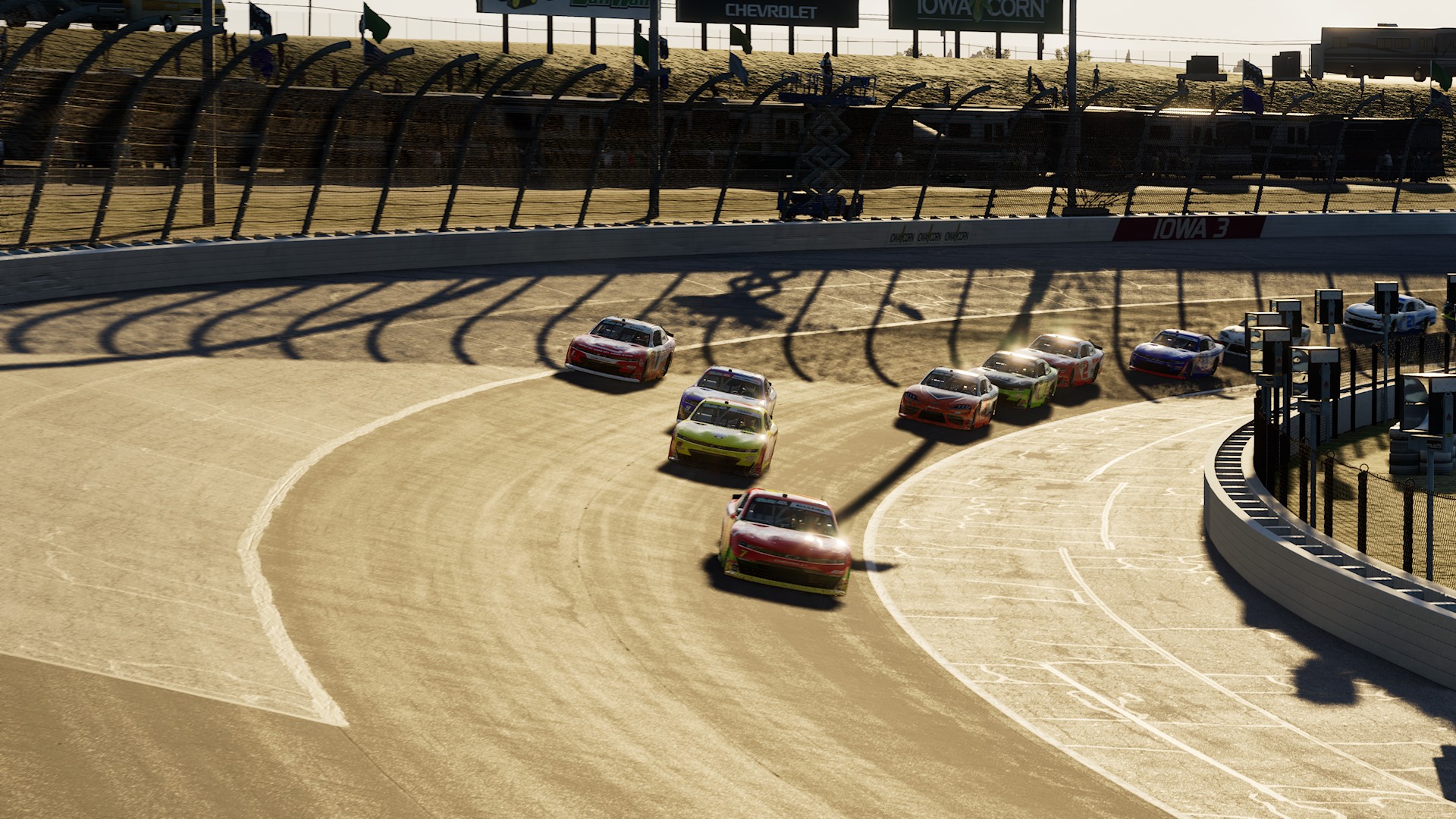 A screenshot from NASCAR 26 showing O'Reilly Series cars racing under a low sun at Iowa Speedway.