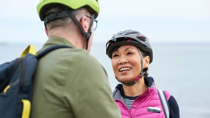 An older couple stands in front of the ocean in bike helmets. The man's back is to us, but the woman faces him, smiling. They look fit and happy.