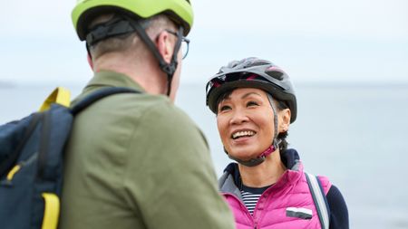An older couple stands in front of the ocean in bike helmets. The man's back is to us, but the woman faces him, smiling. They look fit and happy.