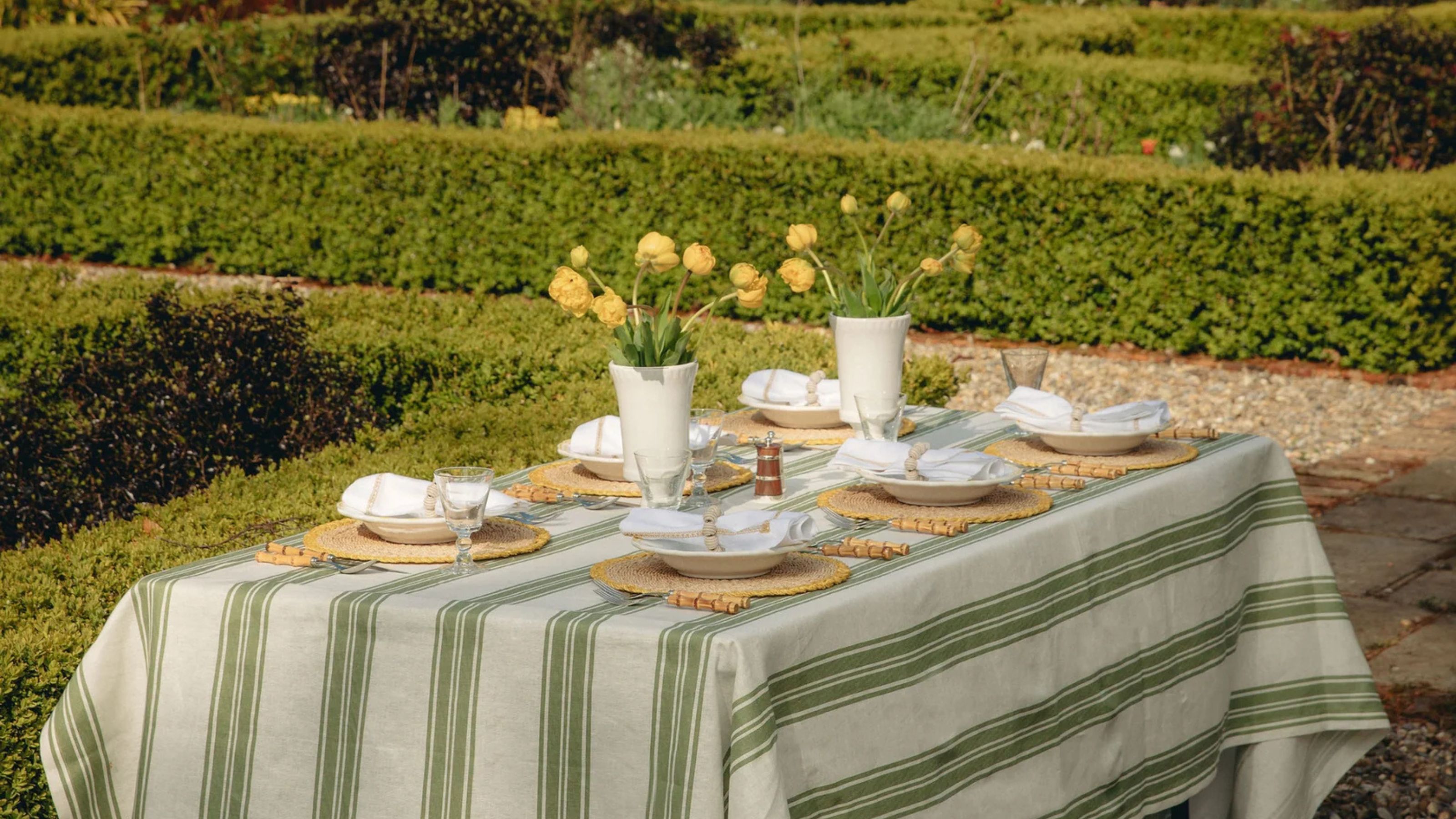 Dinner table in a garden with low box hedges, dressed in a green striped table cloth, woven placemats, bamboo cutlery, white napkins, white bowls, two white vases with yellow flowers, and timber salt mill.