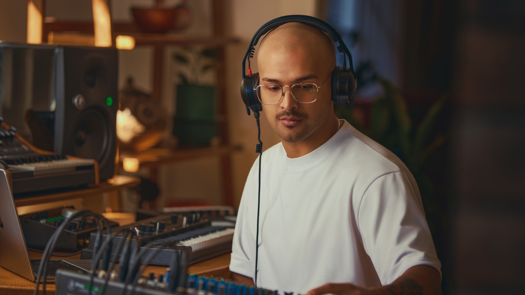 A man in a home recording studio listening to something on a pair of RØDE NTH-50 headphones. The man is wearing glasses and a white t-shirt.
