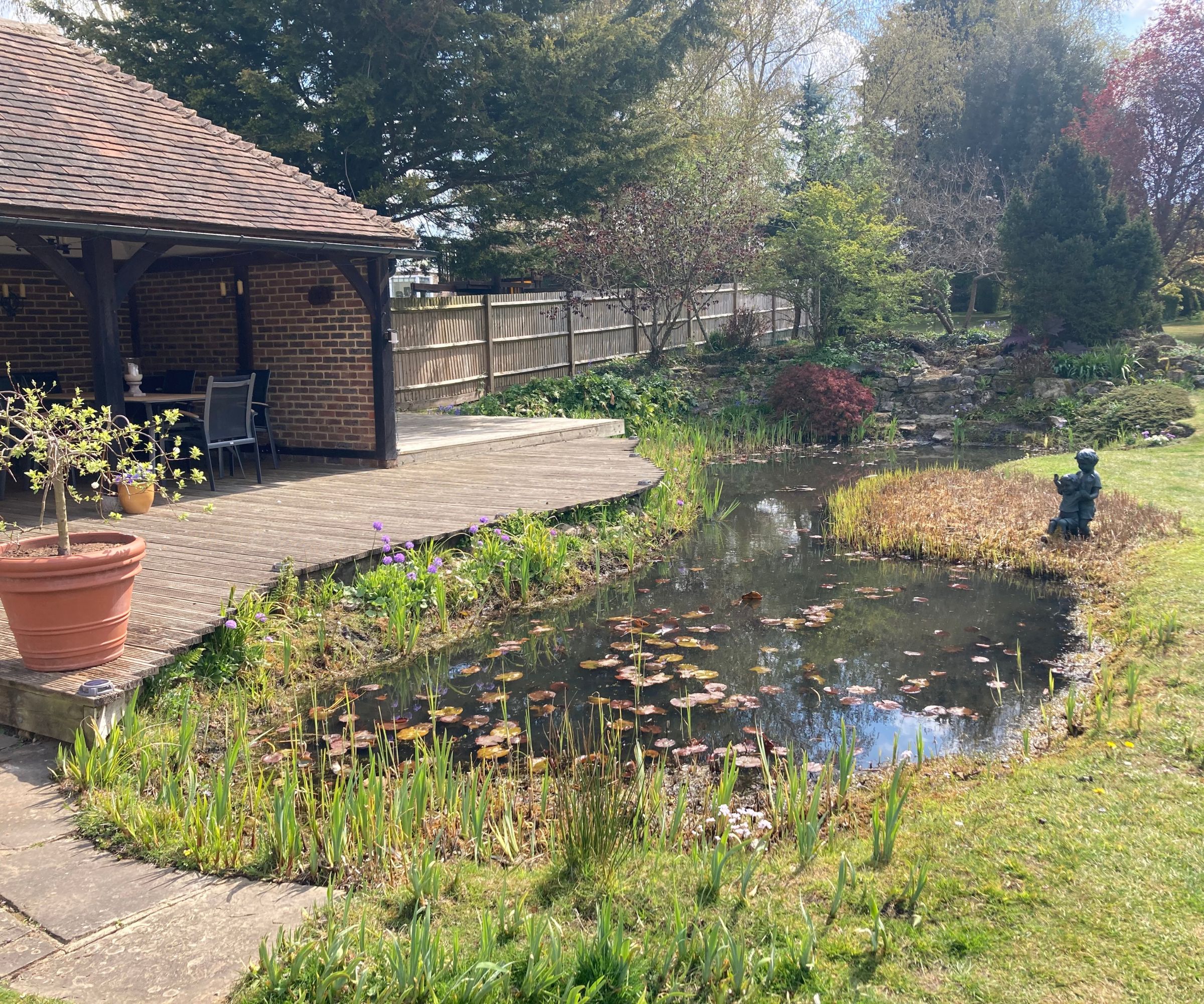 Shady garden pond with lily pads beside a wooden deck and brick pavilion, framed by spring greenery and a peaceful, well-kept lawn.