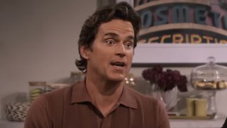 Matt Bomer, sporting longer hair and an open brown collared shirt, is seen in discussion at a table during a scene in Hulu's sitcom, Mid-Century Modern.