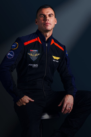 A man stands with arms crossed, wearing dark blue jumpsuits with red accented shoulder wings. He looks serious.