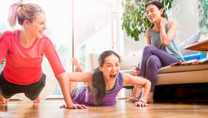 Three women in a living room. The women in the center is lying face down on the floor trying to push herself up, her mouth is open in mock effort. The woman to her left is in the top of a push-up and looks on smiling. The woman to the right sits on a sofa and looks on smiling.