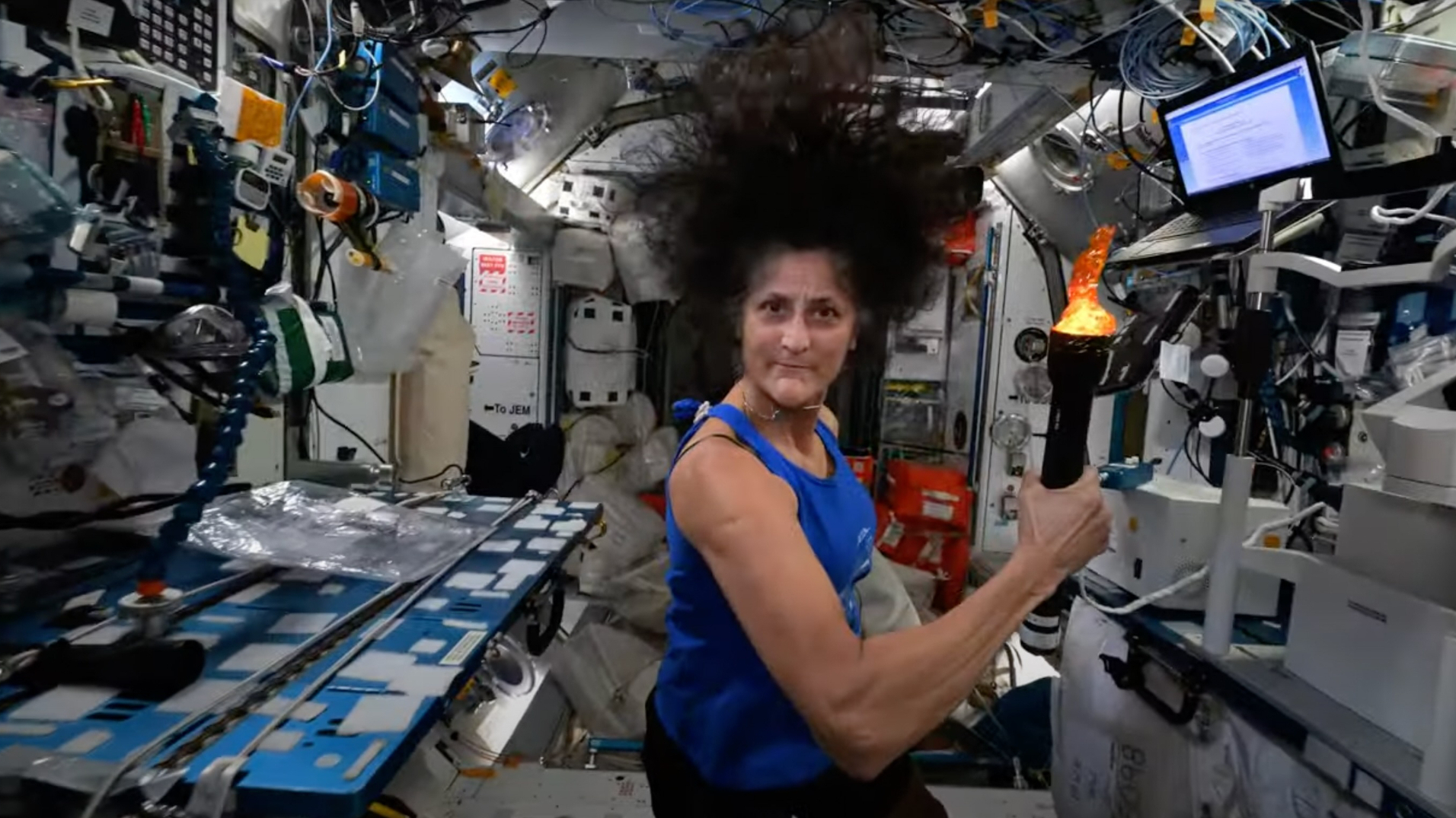 a dark-haired woman in a blue tank top holds a small mock torch aboard the international space station