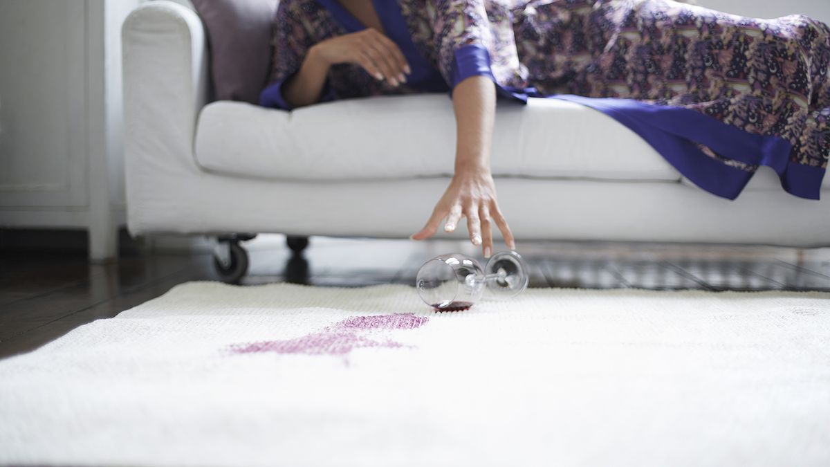 Person reaching down to catch a wine glass from the sofa as it spills on cream carpet