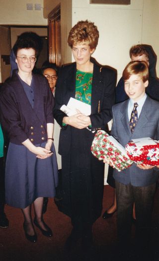 Princess Diana standing with a nun and Prince William, who is wearing a suit and holding Christmas gifts during a visit to homeless charity The Passage