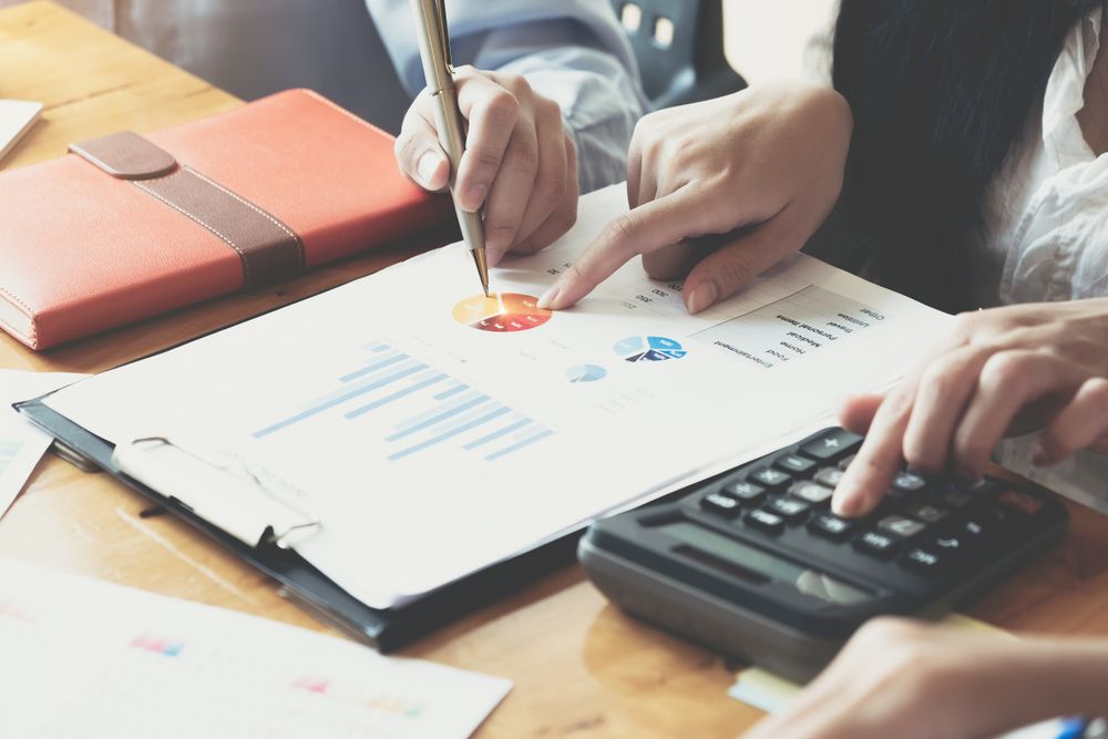 A close up of two people&amp;#039;s hands, pointing to charts on a budget sheet and a third pair of hands doing sums on a calculator