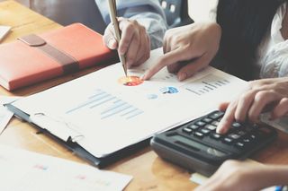 A close up of two people's hands, pointing to charts on a budget sheet and a third pair of hands doing sums on a calculator