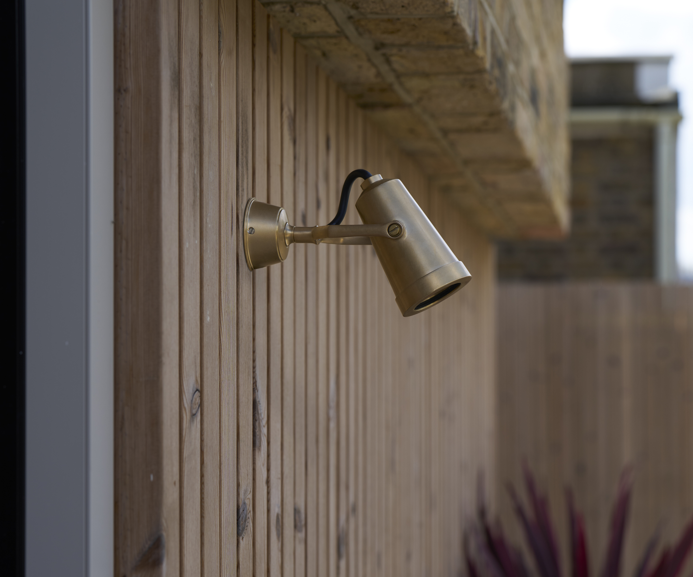 small aged brass finish directional angled spot light on wooden cladded wall below narrow brick overhang