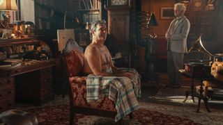 Jon Hamm with a naked chest and a check blanket over his knees sits in an armchair while Michael Sheen in a cream suit as Aziraphale looks on in Good Omens season 2.
