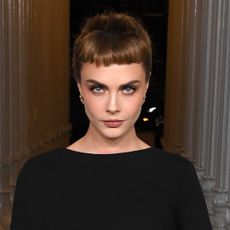 Cara Delevigne with an updo hairstyle that has bangs. She's also wearing a black dress.