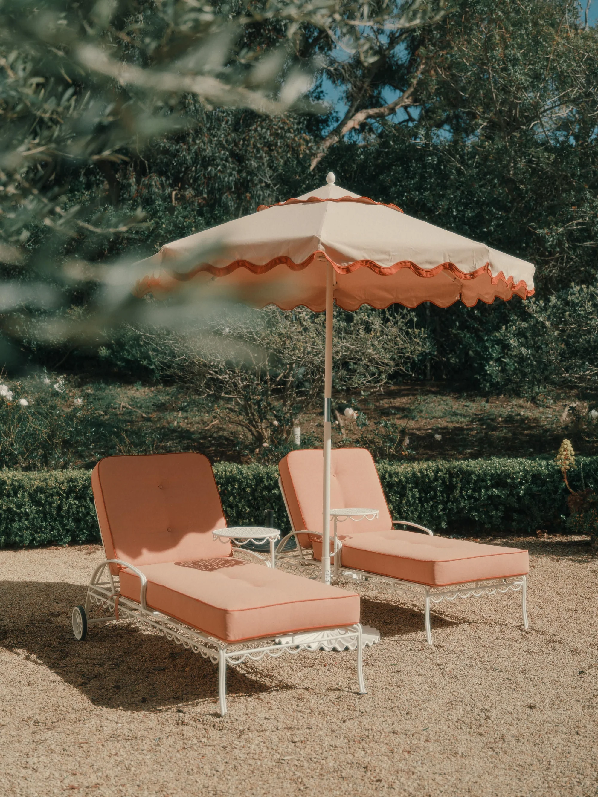 Two coral-pink outdoor loungers beneath a scalloped patio umbrella in a sunlit garden setting