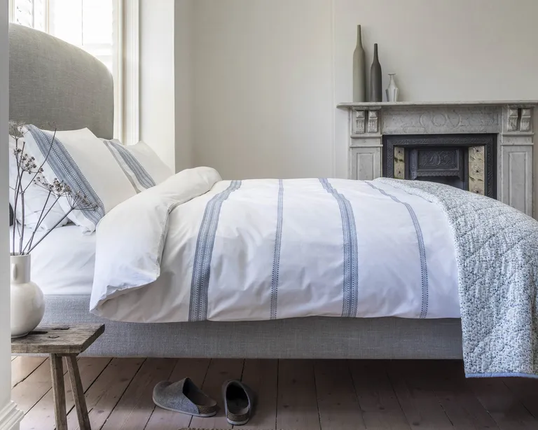 A bedroom with a grey marble fireplace, grey upholstered headboard and white and blue bed linen
