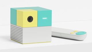 Nex Playground; a small grey, green and yellow cube on a white desk, with a slim controller