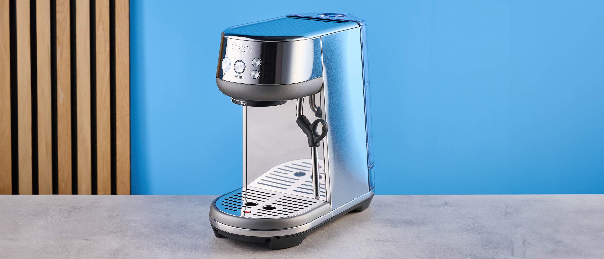 the breville bambino / sage bambino in silver photographed against a blue background with a wooden feature wall, showing the steam wand, group head, water tank, button controls, and espresso