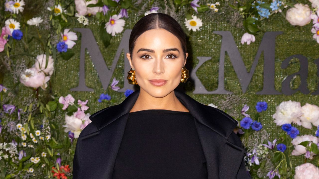 Olivia Culpo wears a black dress, black coat, and has her hair slicked back