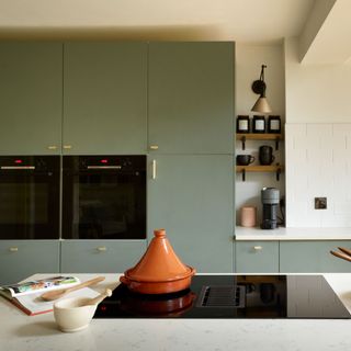 Green/blue cupboards, gold leaf island unit, large patterned tiles above the sink. Wooden dining table, with wooden slatted wall behind. Utility behind dining table and snug area behind kitchen