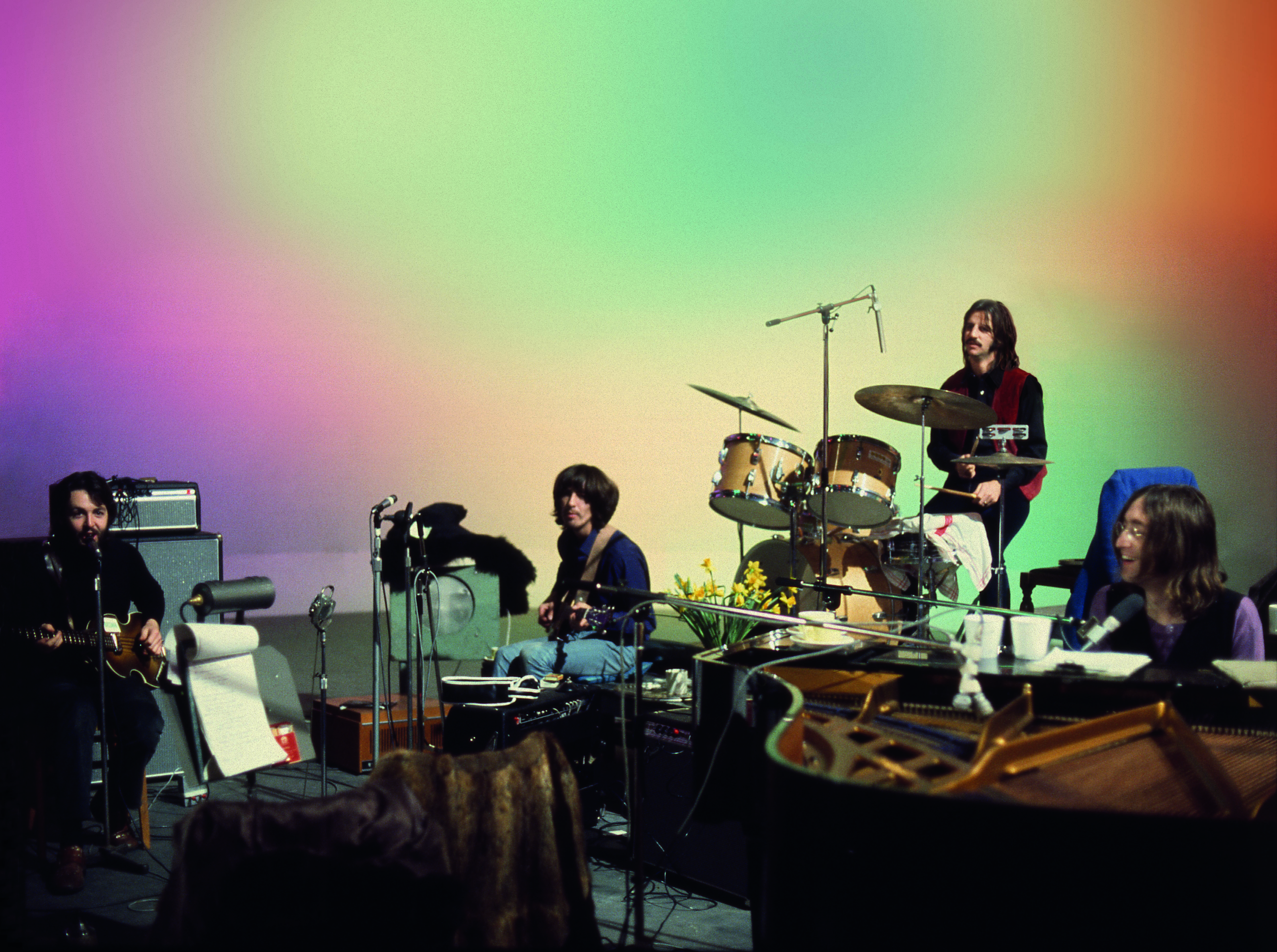 En promobild för The Beatles Get Back där hela gänget sitter och spelar vid sin musikutrustning mot en färgglad bakgrund