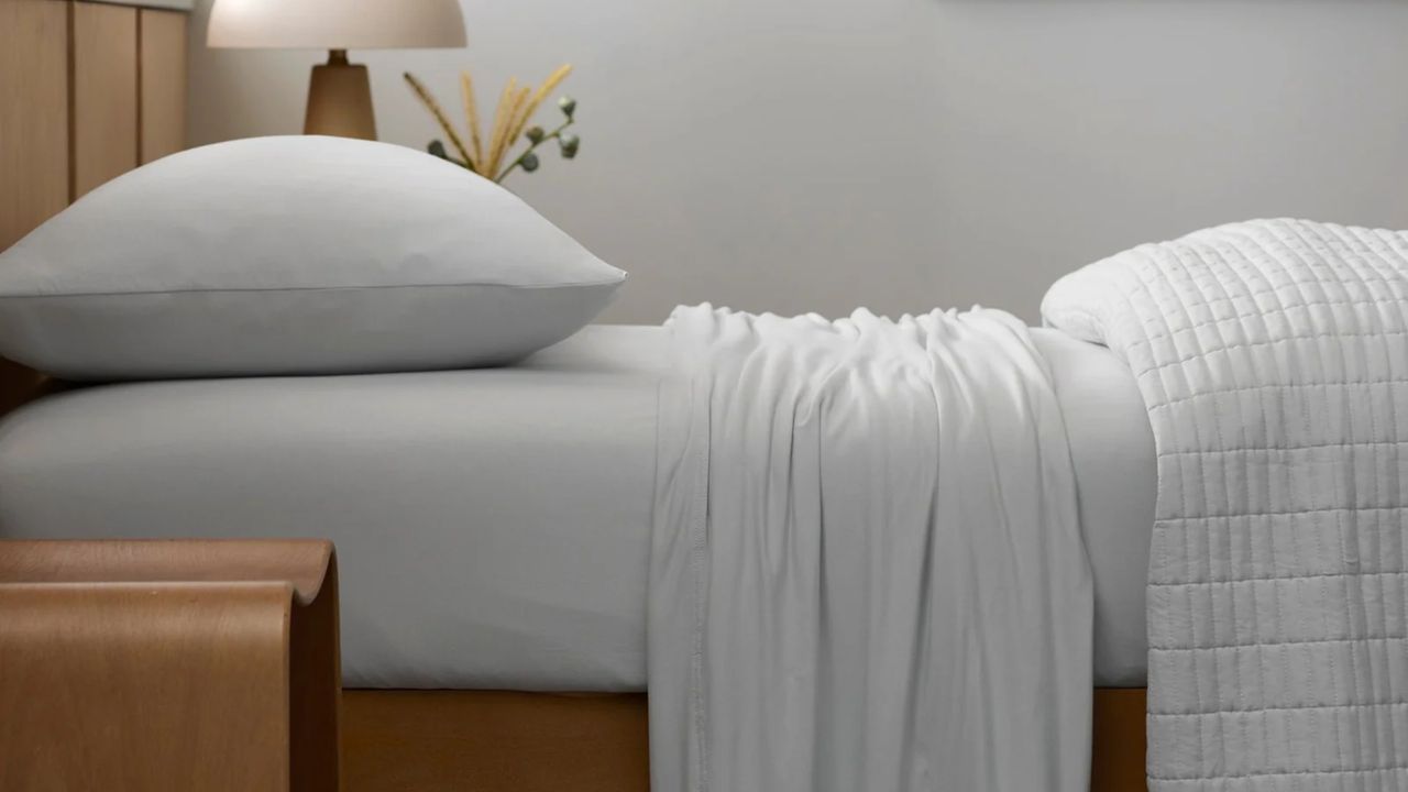 Close-up image of a bed that is made with all-white jersey sheets. There is a quilt folded at the end of the bed and a wooden bedside table with a mushroom lamp.