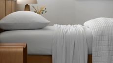 Close-up image of a bed that is made with all-white jersey sheets. There is a quilt folded at the end of the bed and a wooden bedside table with a mushroom lamp.