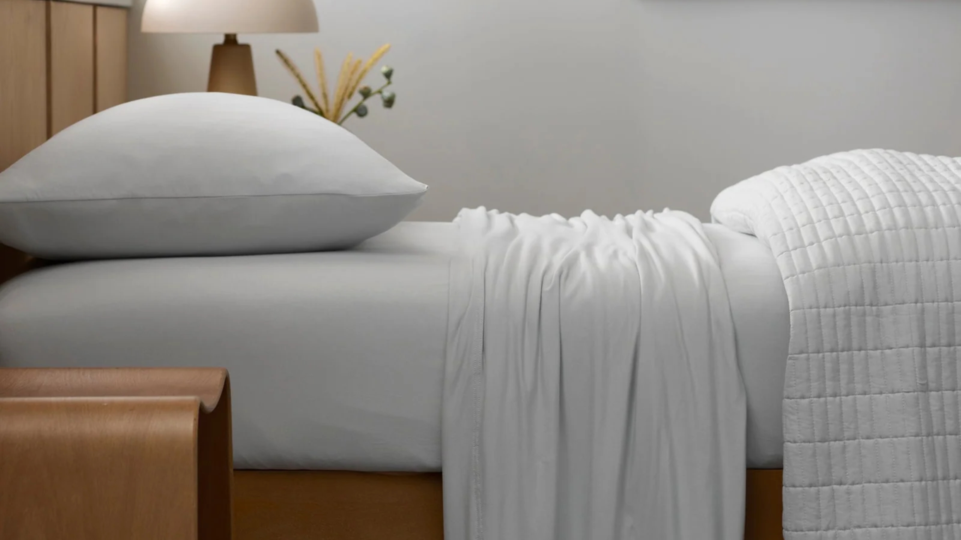 Close-up image of a bed that is made with all-white jersey sheets. There is a quilt folded at the end of the bed and a wooden bedside table with a mushroom lamp.
