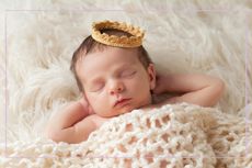 Newborn baby boy wearing a gold crown. He is sleeping on a beige flokati rug with his hands behind his head