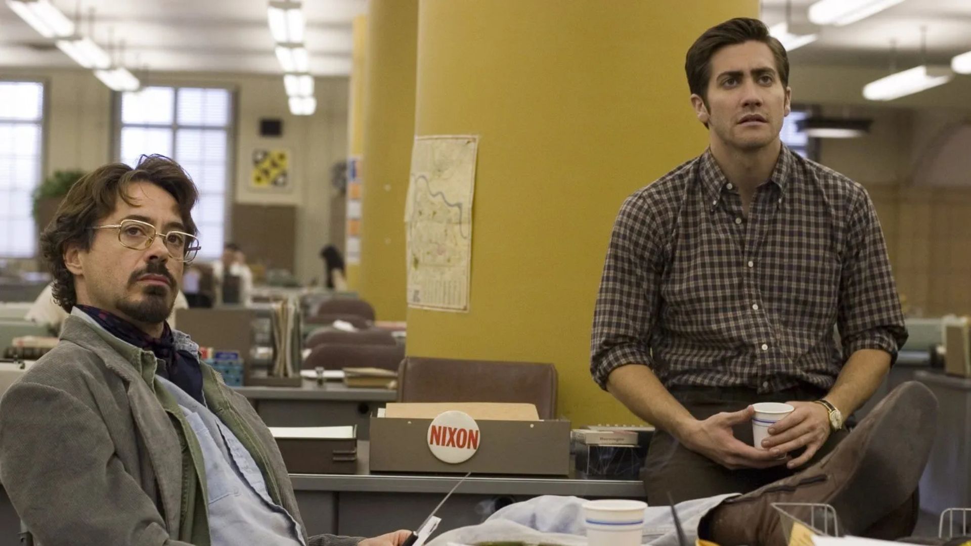 The cast of Zodiac sit on office furniture in the middle of a newsroom