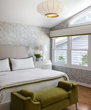 A bright bathroom with large windows, patterned wallpaper, and a green velvet storage ottoman