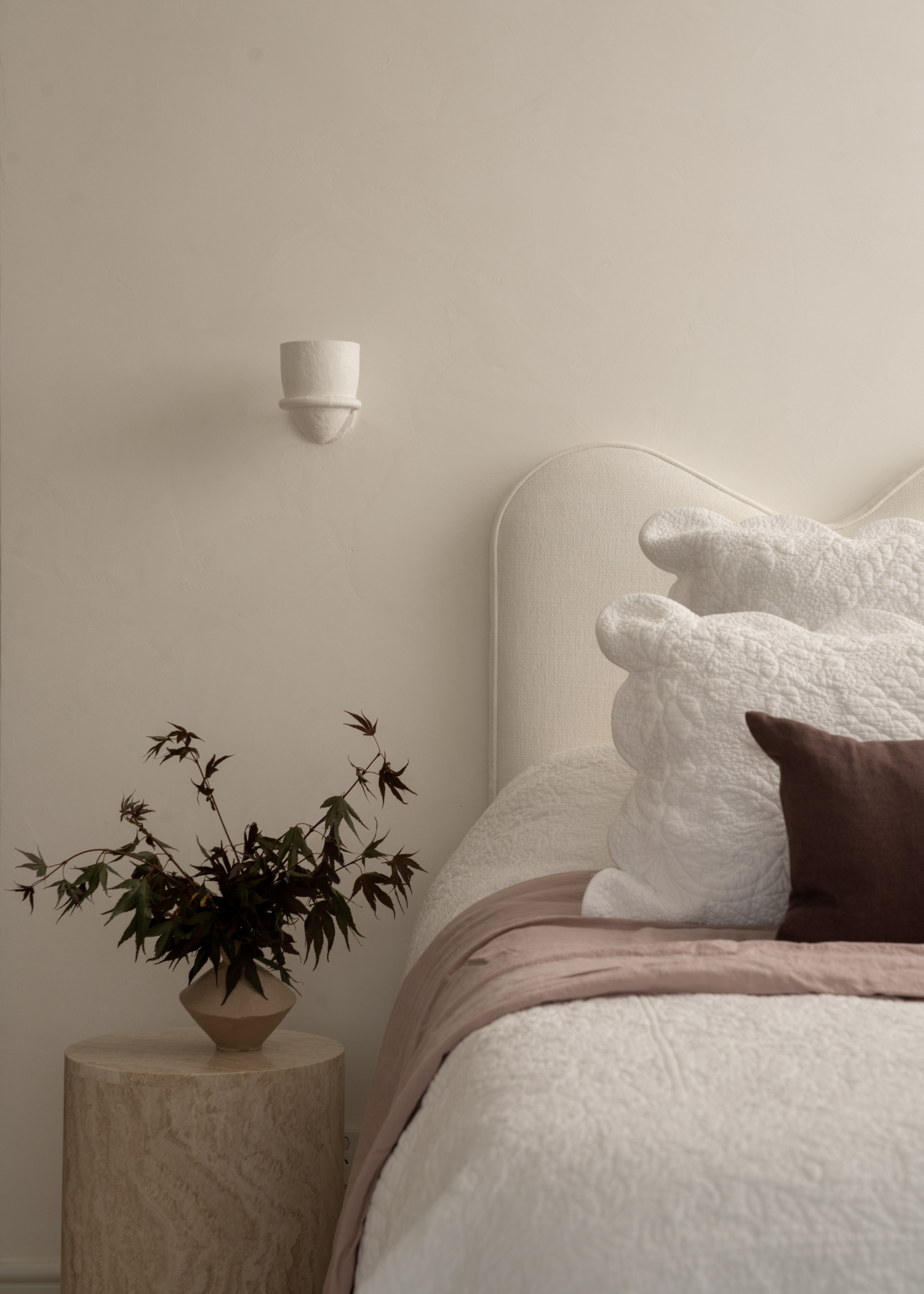 A white bedroom with a wall sconce, a curved headboard, a side stone table with a vase of stems, and textured pillows