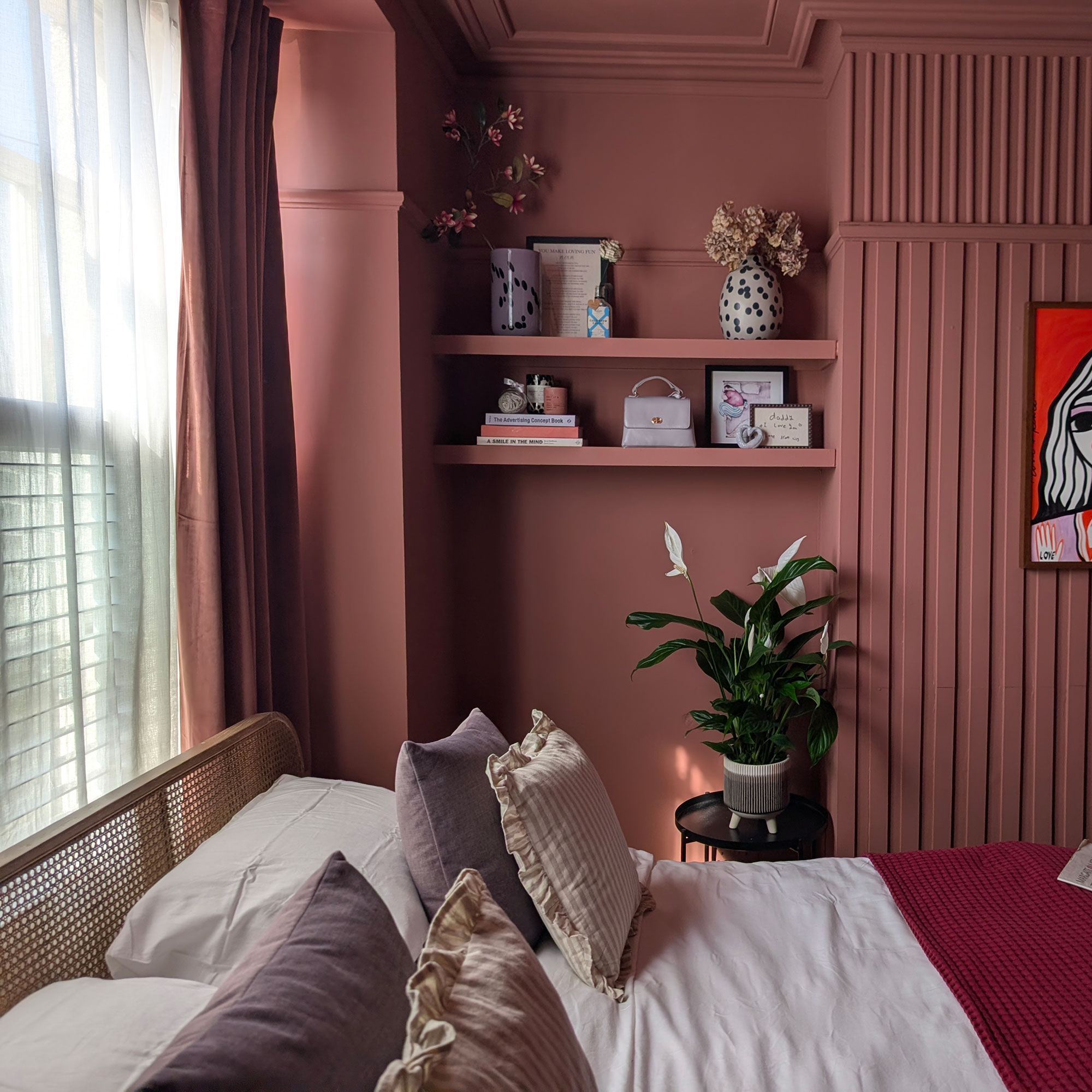 Pink bedroom with wall panelling, floating shelves, rattan headboard and windows layered with sheer and heavy pink curtains