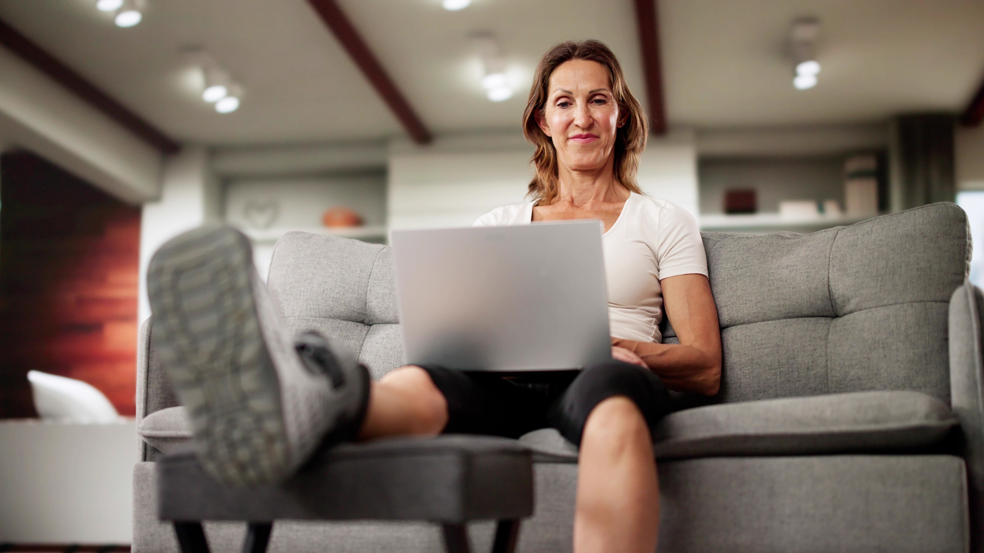 A woman looks at her laptop on her sofa while her leg, in an ankle brace, is propped on an ottoman.