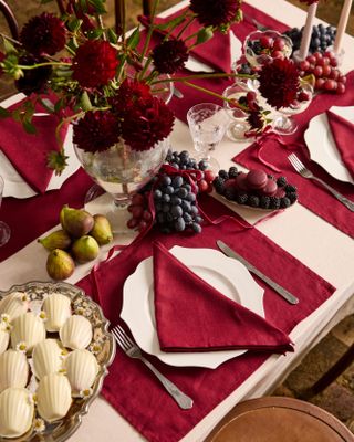 Christmas table setting with a white table cloth, red placemats and napkins, and white plates. There is fruit like grapes and pears along the table and a red floral centerpiece.