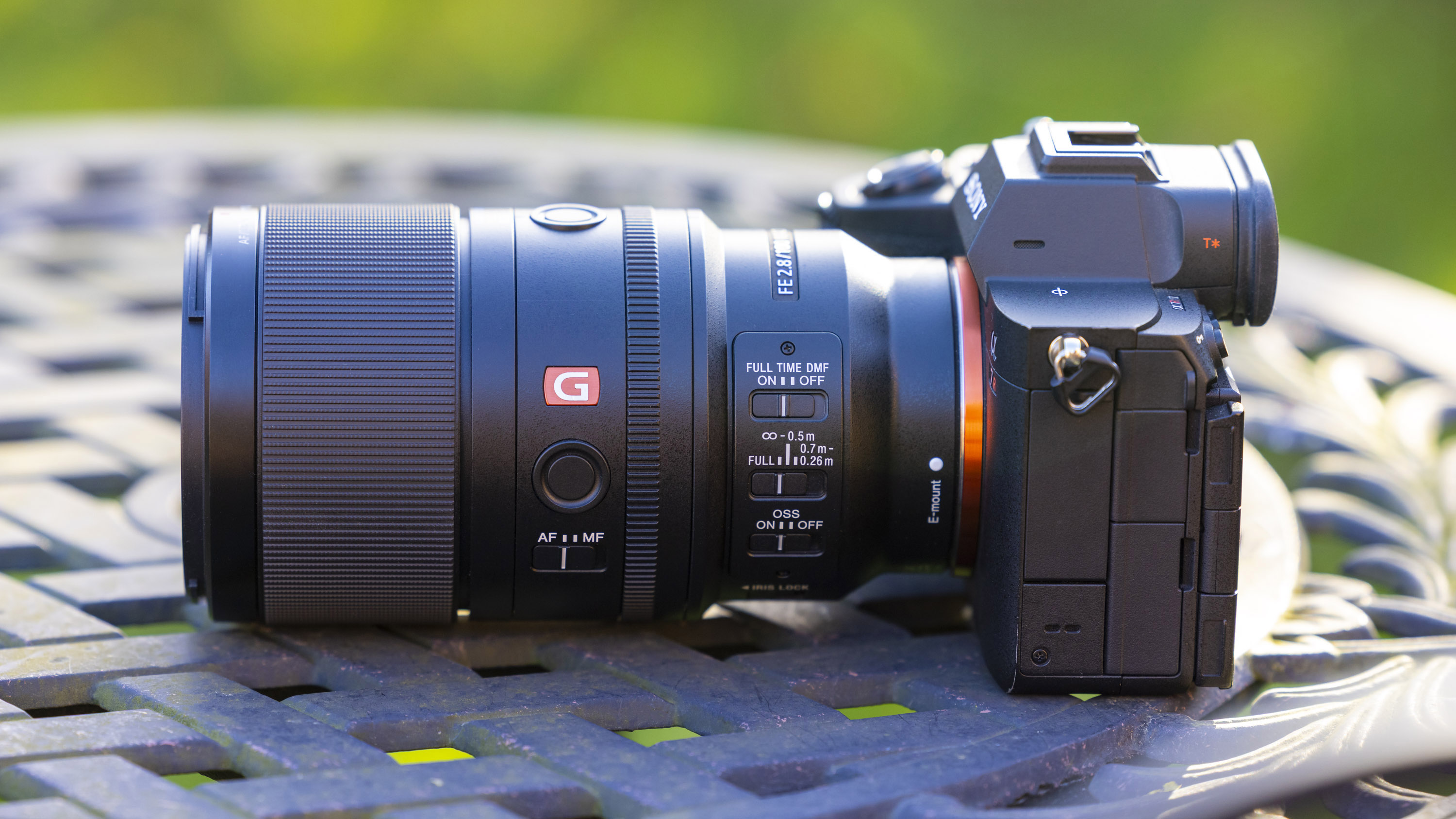 A product shot of the Sony FE 100mm f/2.8 Macro GM OSS attached to a Sony A7R IV outdoors on a metal garden table with a blurry background