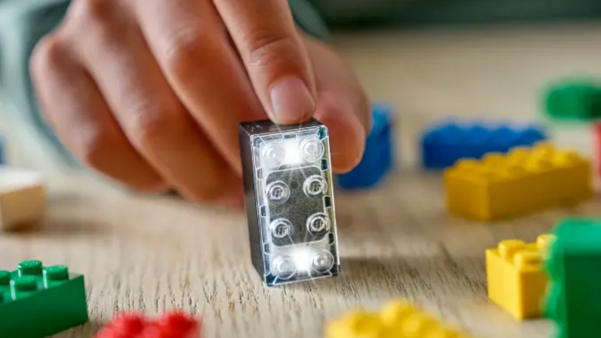 A hand holds the Lego Smart Brick on a table amongst other, normal Lego bricks