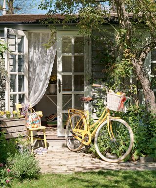 A grey and white painted garden shed with linen curtains, with a yellow bike in dappled shade
