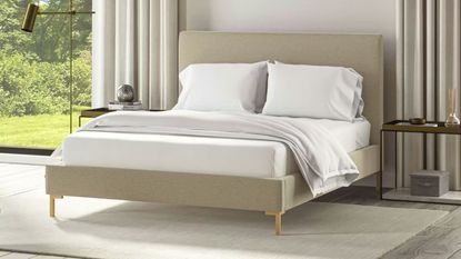 A Saatva bed in a large bedroom with a grey cirrus rug beneath it. To either side of the bed are large windows, with the one to the left visible, with white curtains. The bed is made up with white bedding, and to either side are gold bedside tables.