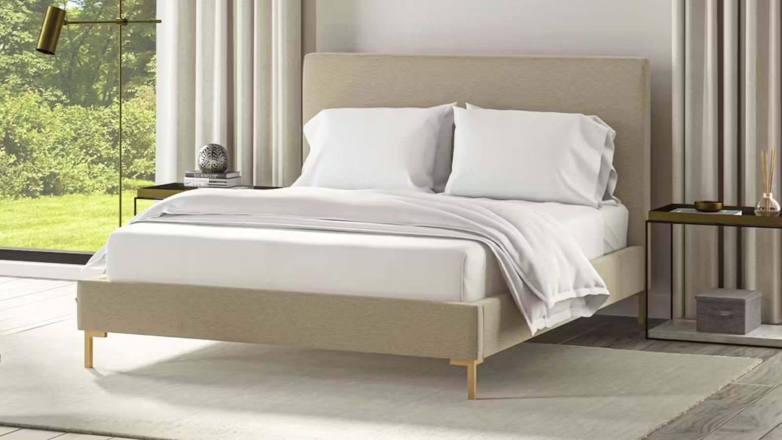 A Saatva bed in a large bedroom with a grey cirrus rug beneath it. To either side of the bed are large windows, with the one to the left visible, with white curtains. The bed is made up with white bedding, and to either side are gold bedside tables.
