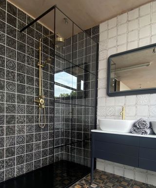 A dark bathroom with black mosaic tiles, a glass shower wall with a brushed gold faucet on it, and a wall with white tiles and a black basin with a mirror with curved edges