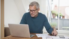 An older man works on paperwork while looking at his laptop at the kitchen table. 